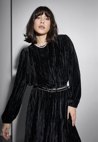 Rochie din catifea neagră cu mâneci lungi, suprafață texturată, strânsă la talie cu o curea neagră accentuată cu argint; un colier cu perle împodobește decolteul.