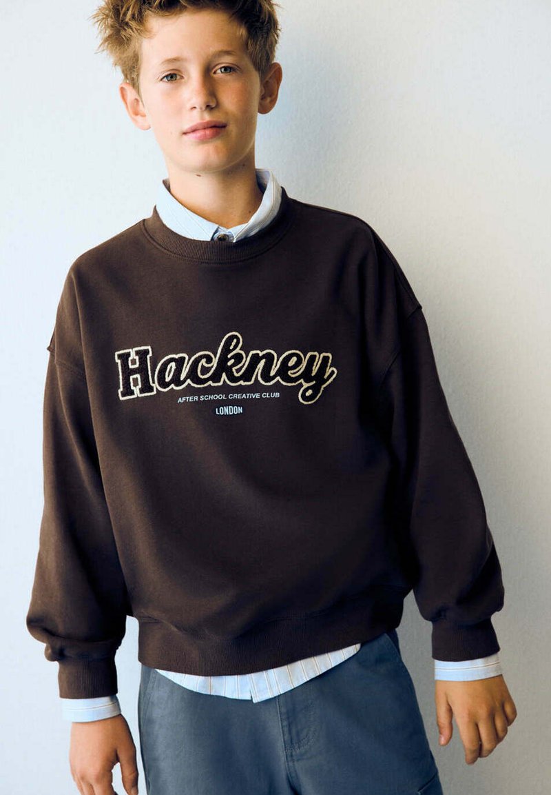 Garçon aux cheveux courts et bouclés portant un sweat-shirt sombre avec le texte "Hackney After School Creative Club London" sur une chemise claire et un pantalon bleu.