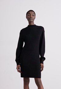 Vestido preto de malha com mangas longas, gola alta e silhueta justa. O tecido texturizado com detalhes canelados confere uma aparência elegante.