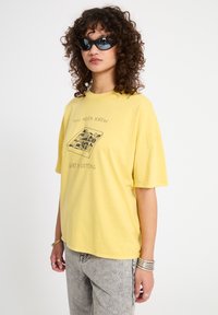 Femme aux cheveux bouclés portant des lunettes de soleil noires, un t-shirt jaune avec un motif et du texte, un jean gris et des bracelets argentés, debout devant un fond blanc.
