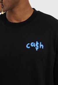 Juodas medvilninis marškinėliai su mėlyna nupiešta "cash" logotipu priekyje. Apvalaus kaklo dizainas su trumpomis rankovėmis, lygi tekstūra.