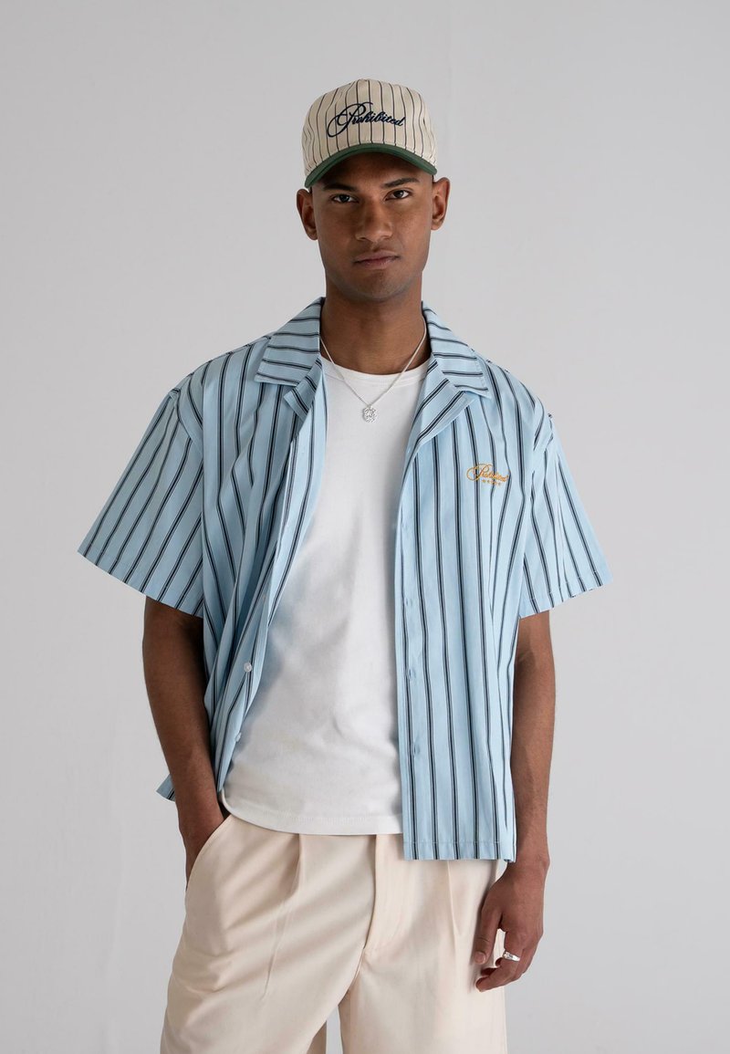Light blue short-sleeve shirt with black vertical stripes, button-up front, and embroidered logo; paired with a beige cap featuring green accents.