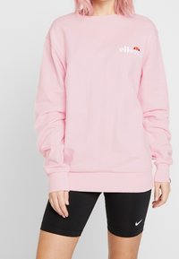 Jasnoróżowy sweatshirt z długimi rękawami i ściągaczem na dole. Posiada haftowane logo na piersi. Noszony z czarnymi szortami rowerowymi.