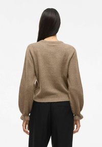 Femme aux longs cheveux noirs portant un pull en tricot marron et un pantalon noir, se tenant de dos contre un fond blanc uni.