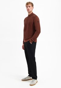 Homme debout portant un pull brun à fermeture éclair quart, un pantalon noir et des baskets Adidas gris clair avec des rayures blanches, regardant droit devant.