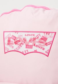 Roza platnena obliž z belo oznako, na kateri je rdeče besedilo "Levi's" in "501" v ukrivljenem, logotipnem izrezu. Obrobljen z roza šivanjem.