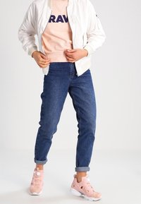 Bílá bomber bunda, pastelově růžový svetr s textem "BRAV", modré zúžené džíny a růžové tenisky s bílými podrážkami a ozdobnými knoflíky.