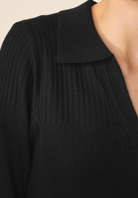 Maglione nero lavorato a maglia con texture a costine, dotato di colletto piatto e scollo a V. Il materiale appare morbido e aderente.
