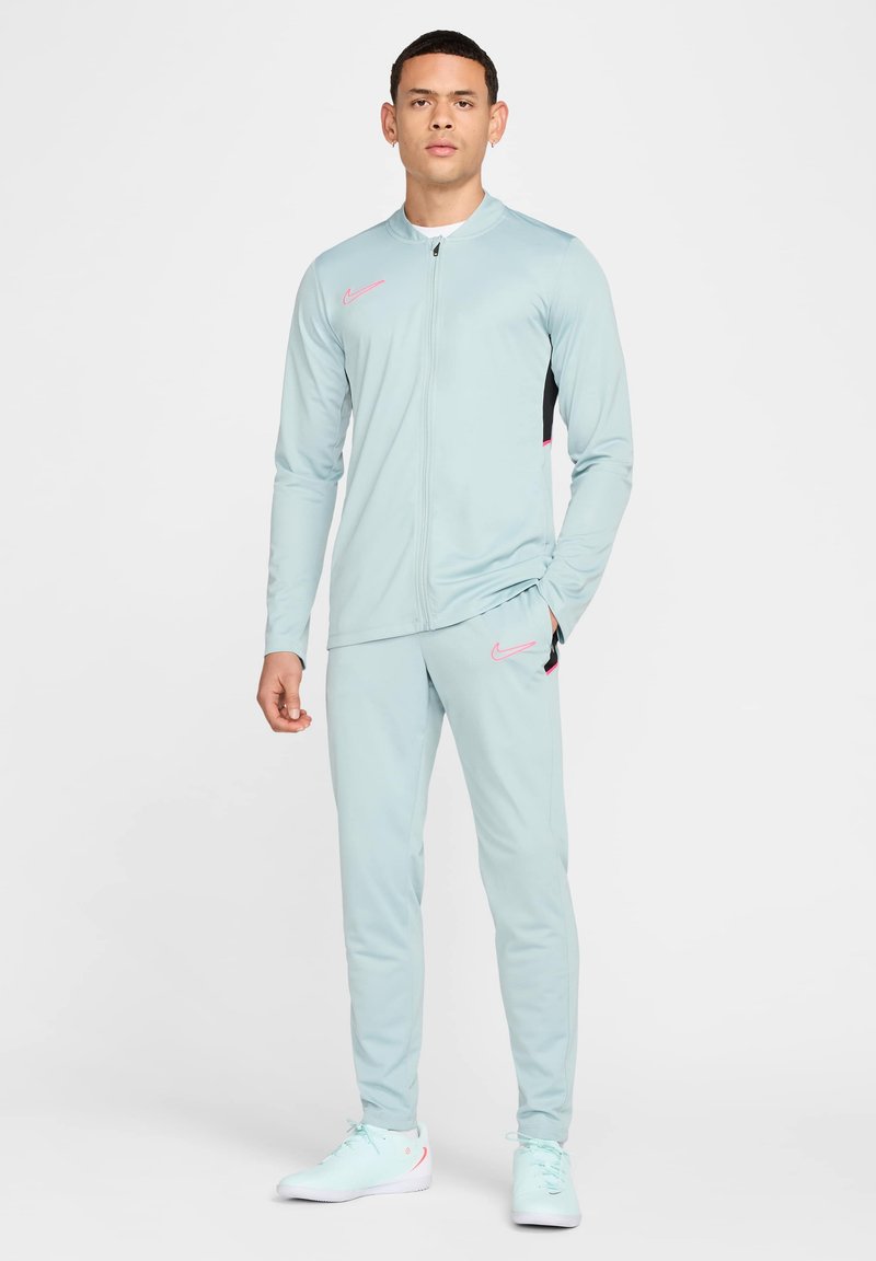 Survêtement Nike bleu clair avec une veste zippée et un pantalon, présentant un panneau noir et des accents roses. Tissu lisse et athlétique, design ajusté.
