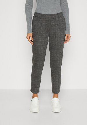 Person trägt graue karierte, zulaufende Hose, graues Langarmshirt und weiße Sneaker und steht vor einem einfarbigen Hintergrund.