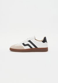 White sneaker with black stripe accents, suede toe cap, and brown rubber sole. Smooth leather upper with a rounded design.