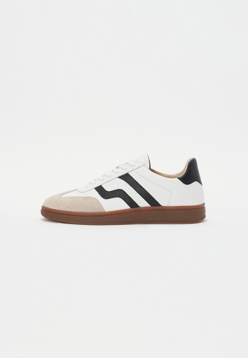 White sneaker with black stripe accents, suede toe cap, and brown rubber sole. Smooth leather upper with a rounded design.