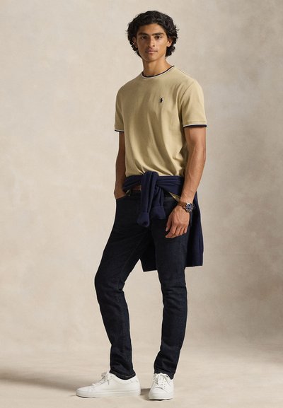 Ανοιχτό κίτρινο μπλουζάκι με ναυτικό γαλάζιο τελείωμα, συνδυασμένο με σκούρο τζιν τζιν. Ένα ναυτικό πουλόβερ είναι δεμένο στη μέση και λευκά αθλητικά παπούτσια ολοκληρώνουν την εμφάνιση.