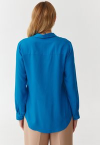 Kvinde med langt blond hår iført en lys blå langærmet skjorte og beige bukser, vendt væk mod en ensfarvet hvid baggrund.