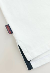Bílé bavlněné tričko v rohu s námořnickým akcentem a červenou nálepkou s nápisem "SUPERDRY." Hladký povrch a prošité lemy.