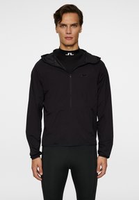 Svart lättviktsjacka med huva och en kvartsydd dragkedja, med en diskret logotyp på bröstet och elastiska muddarmar. Mjuk textur och sportig design.
