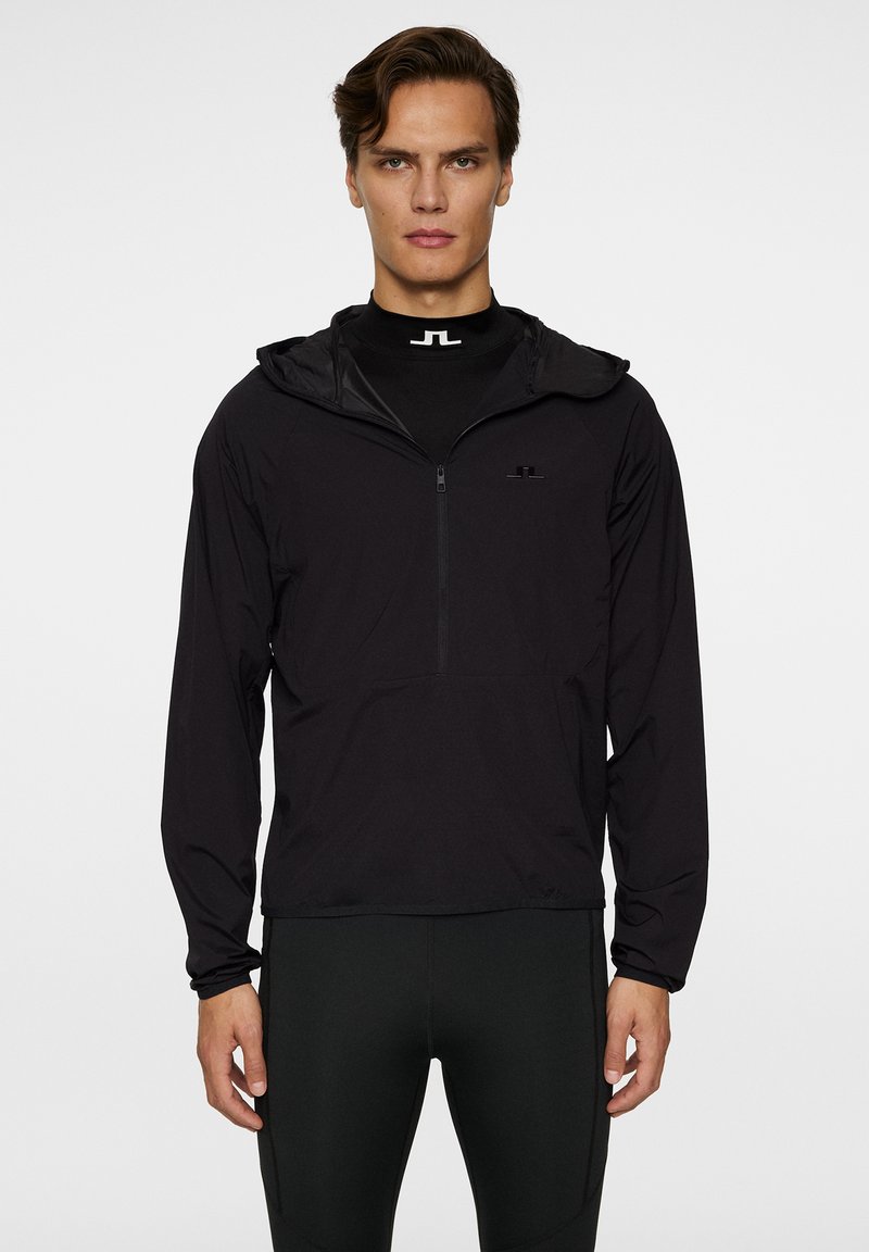 Svart lättviktsjacka med huva och en kvartsydd dragkedja, med en diskret logotyp på bröstet och elastiska muddarmar. Mjuk textur och sportig design.