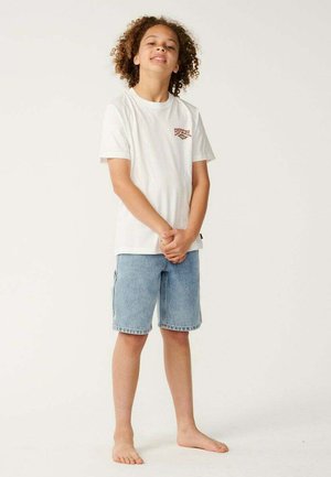 Niño con cabello rizado está descalzo, vistiendo una camiseta blanca y pantalones cortos azules de mezclilla frente a un fondo claro y sencillo.