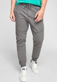 Man i grå drajoggers och vita sneakers med vita strumpor, stående framför en neutral bakgrund.