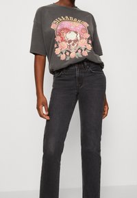 Persona con una camiseta gráfica de color gris oscuro que presenta una calavera, un sol, planetas y rosas rosas, combinada con jeans negros de cintura alta, frente a un fondo simple.