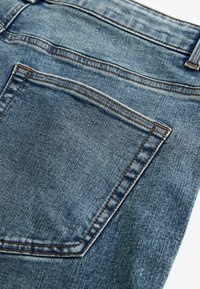 Džínsy z denimu s svetlomodrým umyvaním, s jedným zadným vreckom a kontrastným oranžovým šitím pozdĺž švov a okrajov vreciek.
