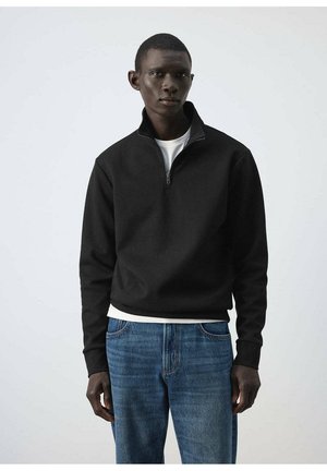 Young man wearing a black quarter-zip pullover over a white shirt and blue jeans, standing against a plain light background.