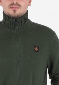Groene zip-up trui met een hoge geribbelde kraag, voorzien van een klein zwart en gouden logo patch op de borst. Zachte, gestructureerde brei-stof.