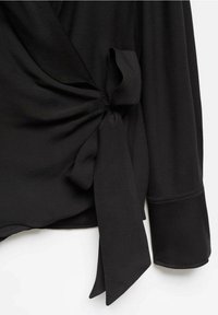 Vêtement noir à manches longues avec un détail de nœud sur le côté et deux pans lâches qui pendent.