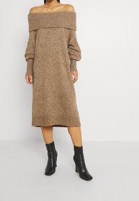 Schulterfreies, langärmeliges Strickkleid in einem hellbraunen Farbton, mit gerippten Akzenten am Ausschnitt und an den Manschetten, kombiniert mit schwarzen Knöchelstiefeln.