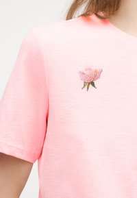 Κοντινό πλάνο ροζ κοντομάνικου πουκάμισου με μικρό σχέδιο ροζέτας από στρας στο στήθος, που φοράει άτομο με ανοιχτόχρωμο δέρμα και καστανά μαλλιά.