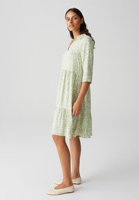 Robe vert clair avec un design à motifs, dotée d'un col, de manches trois-quarts et d'une jupe à volants. Associée à des ballerines beiges.