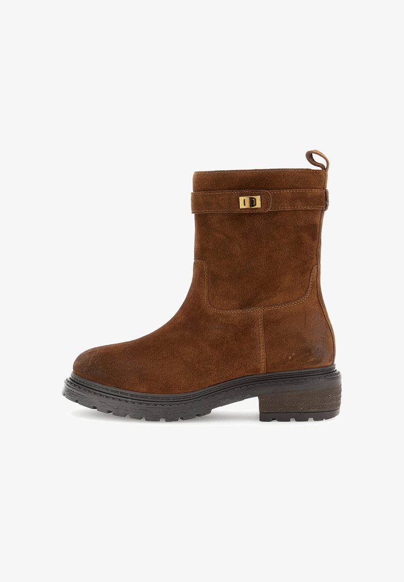 Hnědé semišové kotníkové boty s černou gumovou podrážkou, nízkým blokovým podpatkem, dekoračním popruhem a zlatým přezkou na bílém pozadí.