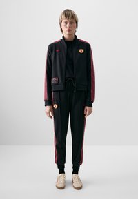 Črn trenirka z rdečimi črtami, blagovno znamko označenimi patche, jakna z zadrgo in hlače z manšeti. Oblečeno z svetlimi čevlji.