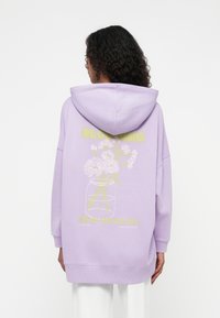 Hoodie lavanda com uma grande impressão gráfica de flores num frasco, apresentando o texto amarelo "BLOOMING NEW-SEASON." Tecido macio, corte descontraído.