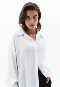 Camisa blanca de gran tamaño con cuello y mangas largas. Textura suave, corte relajado, combinada con un sencillo collar de oro.