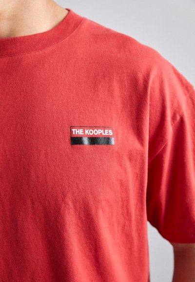 Rød bomulds T-shirt med rund halsudskæring og korte ærmer, med et rektangulært mærke "THE KOOPLES" i hvid og sort på brystet.