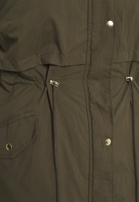 Chaqueta verde oliva con un acabado suave y mate. Cuenta con herrajes dorados, cordones ajustables en la cintura y un bolsillo frontal.