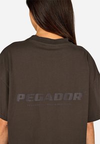 Brązowy bawełniany T-shirt z okrągłym dekoltem i krótkimi rękawami. Na plecach znajduje się wypukłe, ciemne logo z napisem "PEGADOR."