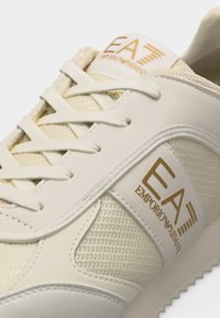 Κοντινό πλάνο κρεμ χρωματιστού αθλητικού παπουτσιού EA Emporio Armani με πλέγμα υφάσματος, δερμάτινες λεπτομέρειες, λευκά κορδόνια και χρυσό λογότυπο στη γλώσσα και το πλαϊνό τμήμα.