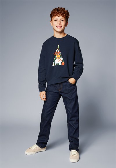 Sudadera de color marino con un gráfico de escena invernal colorido, combinada con jeans de mezclilla oscura y zapatillas ligeras de color beige. Texturas suaves, corte casual.
