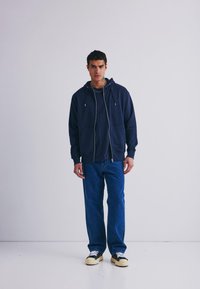 Mornarska zip-up kapuca s vrvicami, v kombinaciji z mornarsko T-majico in modrimi ravnimi hlačami. Obutev vsebuje črne in kremne akcentne barve.