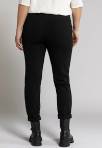 Pantalones negros ajustados de cintura alta con una textura suave y dobladillos enrollados, combinados con botas de combate negras. La vista trasera muestra que no hay bolsillos visibles.