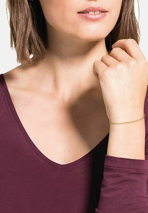Donna che indossa un bracciale d'oro, una maglietta bordeaux a V, con la mano vicino al viso e i capelli corti castani in vista.