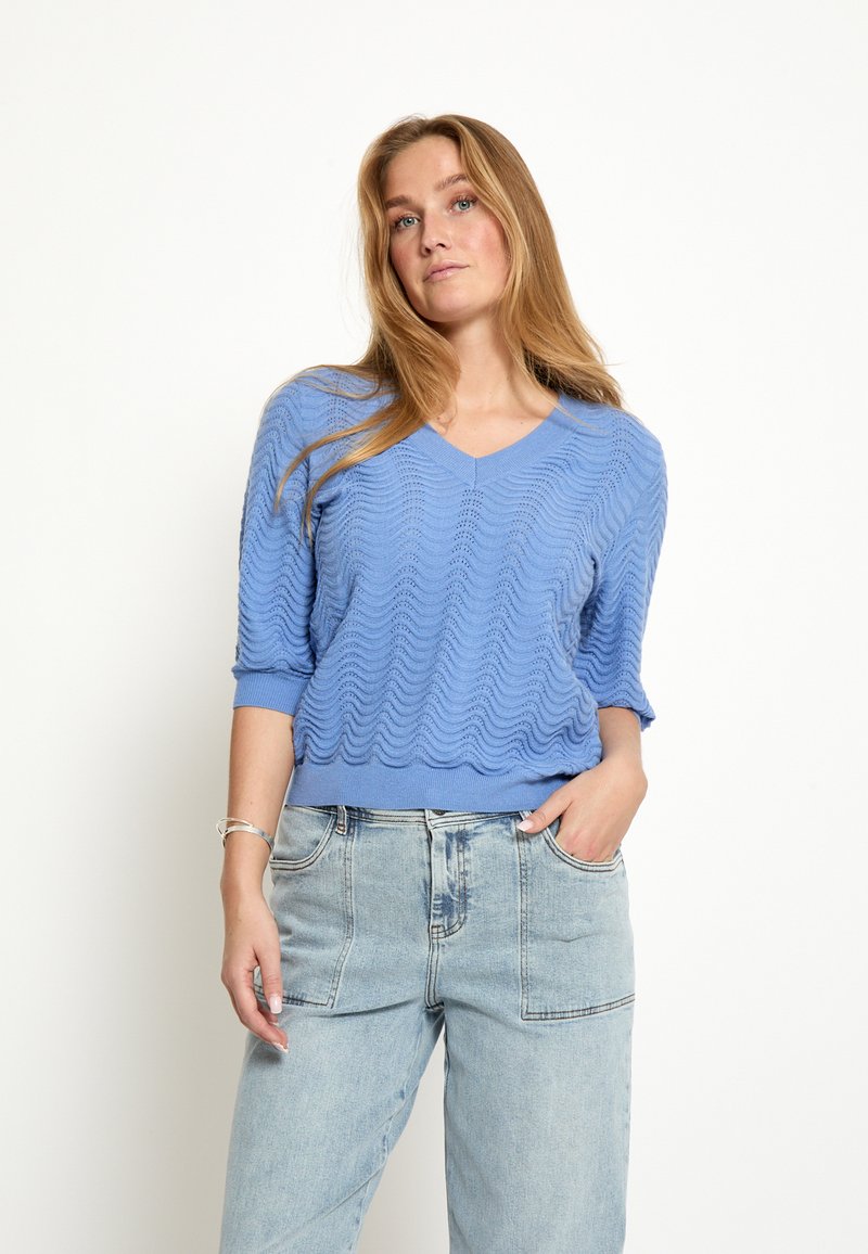 Mujer con un top de punto texturizado azul claro con mangas hasta el codo y jeans de lavado claro, de pie con una mano en el bolsillo.