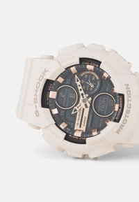 Montre G-Shock avec un boîtier en résine beige, un cadran noir, des accents en or rose, un affichage numérique multifonction et un bracelet en silicone texturé.