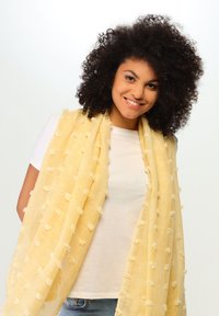 Bufanda amarilla con textura y suaves acentos esponjosos, colocada sobre una camiseta blanca. Tela ligera y transpirable, perfecta para superponer.