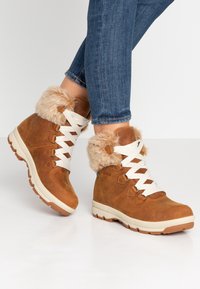 Bruine leren enkellaarzen met pluche beige nep-bont boorden, witte veters en rubberen zolen, voorzien van een gestructureerde afwerking en contrasterende accenten.