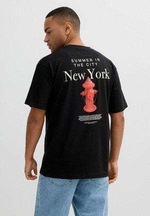 Hombre de espaldas con camiseta negra que lleva el texto "Summer in the City New York" y gráfico de boca de incendio roja, combinado con jeans azul claro.