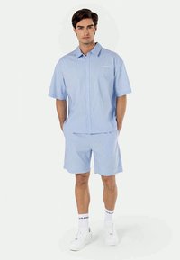 Light blue, short-sleeve shirt and matching shorts with white stripes. Smooth fabric, button-up shirt, and relaxed fit. White sneakers and socks.