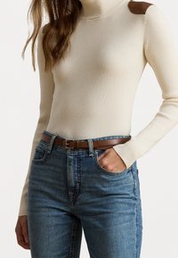 Žena v krémovém žebrovaném roláku s hnědými nášivkami na ramenech, modrých džínách a hnědým koženým pásem, s jednou rukou v kapse.